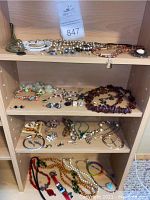 Three shelves containing assorted costume jewelry including beaded necklaces, earrings, rings and a cameo pendant necklace.
