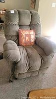 Full frontal view of beige fabric recliner with remote control cord visible, featuring a square patchwork decorative pillow on the seat.