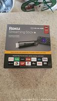 Photo of Roku Streaming Stick+ packaging showing branding, features (HD 4K HDR, voice remote) and supported streaming services
