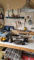 Workbench with assorted hand tools, Ryobi cordless drill and accessories arranged on a white table with pegboard in background