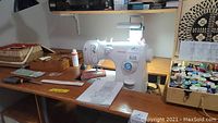 Singer Inspiration sewing machine on a wooden table with assorted sewing supplies and instruction manual spread around it. OTT-Lite lamp is turned on above the sewing machine providing light.