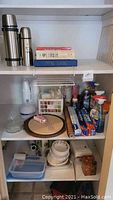 Wide shot of two shelves showing thermoses, cleaning supplies, disposable dishware, recipe books, and miscellaneous kitchen items.