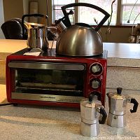 Red Hamilton Beach toaster oven with stainless steel Copco whistling teapot on top, two small aluminum stovetop espresso makers, and small stainless steel pitcher.