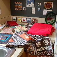 Various art tiles with colorful and natural designs including hummingbirds, cups, sunflower, and abstract patterns displayed on a ledge above the countertop. Below are kitchen linens including a folded bright red tablecloth and patterned cloth towels and napkins in earthy tones.