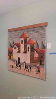Full view of the woven wall hanging showing village scene with buildings, human figures, and mountains in earth tone colors with red roofs.