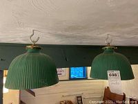 Two green ribbed metal shades hanging side by side from ceiling hooks against a dark beam, one bearing the lot tag visible.