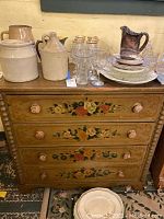 Front view of the chest showing four drawers with floral hand-painted details and round wooden knobs, along with some pottery on top.