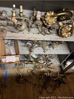 Top shelf of metal shelving holding assorted brass and silver wall sconces in candle-style sockets.