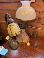 Two desk lamps on wooden table: a gooseneck stained glass lamp and a white milk glass shade student lamp.
