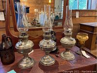 Group view of three stainless steel oil lamps, amber glass growler lamp, and reproduction metal base with amber shade on wooden table