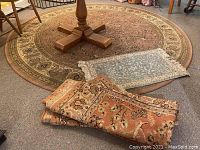 Photo showing large round beige floral rug with part of table base, small blue rectangular rug on top, and rolled large orange floral rug in front.