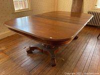 Rectangular solid wood dining table with rounded edges, two carved pedestal legs on casters, polished medium-brown finish, shown in room with wooden floor and wallpapered walls.