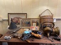 Full view of assorted antiques on a table including framed wall art, brass sconce, wooden bucket, meat grinder, vintage cake decorator and other metal and glass items.