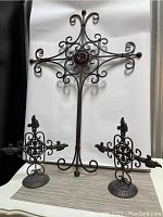 Front view of the large ornate wrought iron cross with a central red flower and two smaller crosses each on round bases.