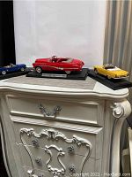 Three collectible model cars displayed on a white dresser, including a red 1949 Buick Roadmaster on a base, flanked by blue and yellow convertible models, likely a 1964 Mustang and 1957 Thunderbird.
