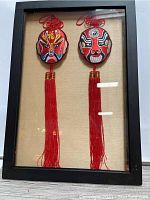 Framed Asian decorative masks with red embroidery and long red tassels against a beige background inside a black frame.