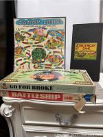 Stacked vintage board game boxes including Battleship with damaged box, Go For Broke with intact box, Uncle Wiggily in black sleeve, and Candy Land board behind.
