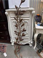 Full view of the wrought iron vine and leaf sculpture standing against a white ornate cabinet.