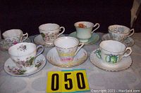 Photo showing 7 assorted tea cups with matching saucers arranged on a table. Various floral and patterned designs are visible across the cups and saucers.