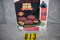 Front of the George Foreman Super Champ Limited Edition grill box showing cooked steaks and key features of the grill including patented grooves and sloped grill, as well as the branding and promotional text.