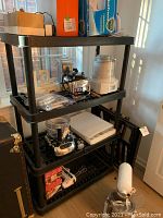 First image shows one black plastic 4-tier shelving set loaded with various small kitchen appliances and items including a blender and food processor.