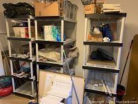 Three white plastic shelving units with five shelves each, holding miscellaneous household items. Shelves have slatted design and black corner supports.