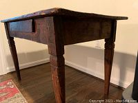 Side angle of antique farmhouse wooden table showing the carved tapered square legs and thick worn top.