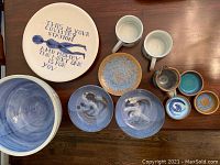 Top view of the full collection showing a large decorative plate with text, mugs, bowls, small plates, and a pitcher predominantly in blue and brown glazes.