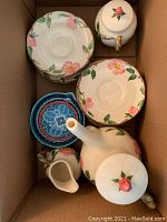 Top-down photo of the box showing coffee pot, creamer, sugar bowl, and several saucers and plates with floral pink rose pattern on cream background.