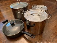 Three large stainless steel pots with matching stainless steel lids and one large black Tfal sauce pan with glass lid, arranged on a wooden floor.