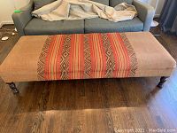 Full view of the ottoman with original striped upholstery and wood legs, positioned in front of a gray sofa.