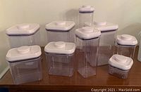 Photo showing 18 clear plastic Oxo food storage containers with varying shapes and sizes, all with white airtight lids arranged on a wooden surface.
