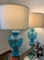 Pair of blue glass ginger jar shaped table lamps on glass surface, showing blue translucent bases and white drum shades.