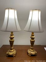 Front view of the pair of gold toned table lamps with white fabric shades on a wooden surface.