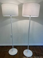 Pair of white floor lamps standing side by side against a white wall, showcasing the full height and design with illuminated shades.