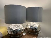 Photo showing two silver mirror glass table lamps side by side with white cylindrical shades on a wooden surface.