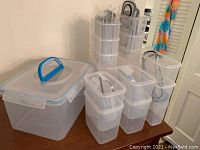 Photo showing a large clear plastic container with a blue handle lid beside stacks of smaller rectangular containers with locking lids.