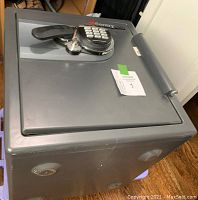 Front-side angled view of a gray metal Sentry Safe showing electronic keypad lock with black handle and round key lock below. The safe is resting on a wooden floor.