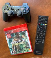 Sony DualShock 3 wireless controller, black color, with classic PlayStation buttons and sticks next to the game case and remote control.