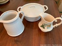 White ceramic serving dish with lid and two ceramic pitchers placed on wooden table.