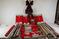Full lot view showing 11 rolls of holiday wrapping paper and various red bows and ribbons spread out on table.