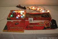 Two vintage original Noma Christmas light boxes with working light sets illuminated. Bundled wires and assorted colored bulbs visible. Boxes show wear and damage.
