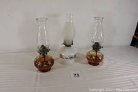 Three kerosene oil lamps shown side by side on a white surface, two amber glass with colorful elves and one white milk glass with winter scene, all with glass chimneys and metal burners.