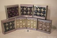Photo showing 7 unopened boxes of glass Christmas ball ornaments arranged to show colors and brands. Colors include various shades of purple, blue, silver, and white. Labels visible for Martha Stewart and Sandra.