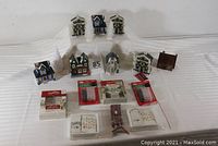 Wide shot showing all eight miniature houses, three white trees, a brown village fence bag, and multiple packaged and unpackaged miniature village lighting items arranged on a white cloth background.