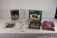 Full lot on table including snow globe kits, hooked rug kits in boxes, books, and ornament storage container.