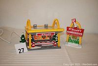 Photo of ceramic McDonald's Snow Village building and separate illuminated sign against a plain background, showing one loose arch on building, holiday decorations, and light cord.