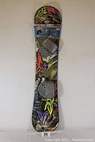 Full view of the snowboard standing upright showing colorful graffiti-style graphics and two attached grey bindings.
