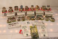 Photograph showing 19 small collectible houses from the Country Cottage and Americana Collections with original boxes and accessory pieces, all arranged on white artificial snow blanket.