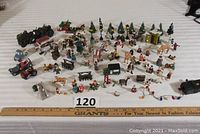 Wide view of the entire collection including the miniature people, trees, light poles, small vehicles, animals, and battery lights arranged on a surface with a ruler showing scale